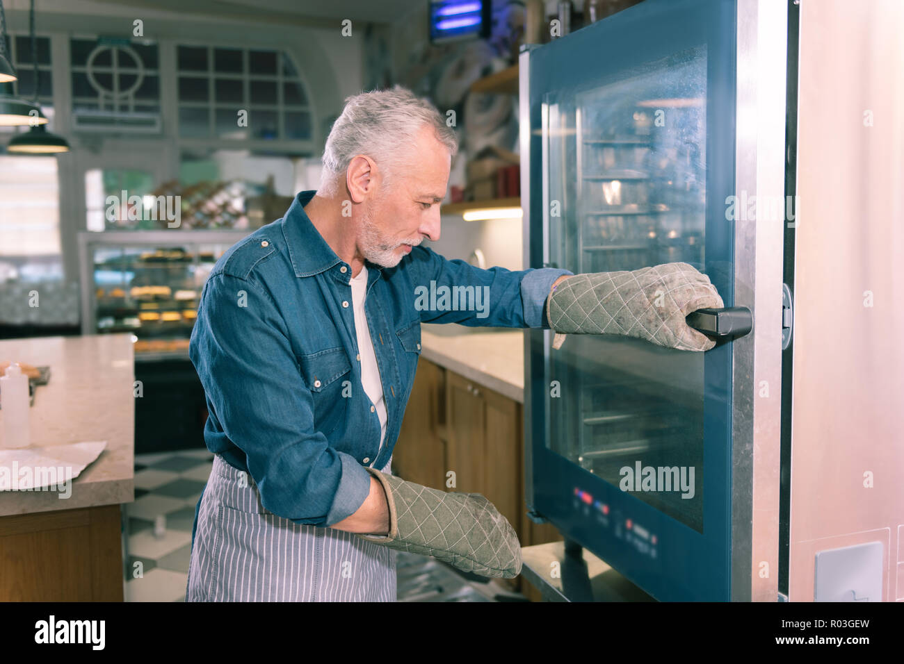 Grey-haired proprietario del panificio indossando il denim shirt in piedi vicino al forno Foto Stock