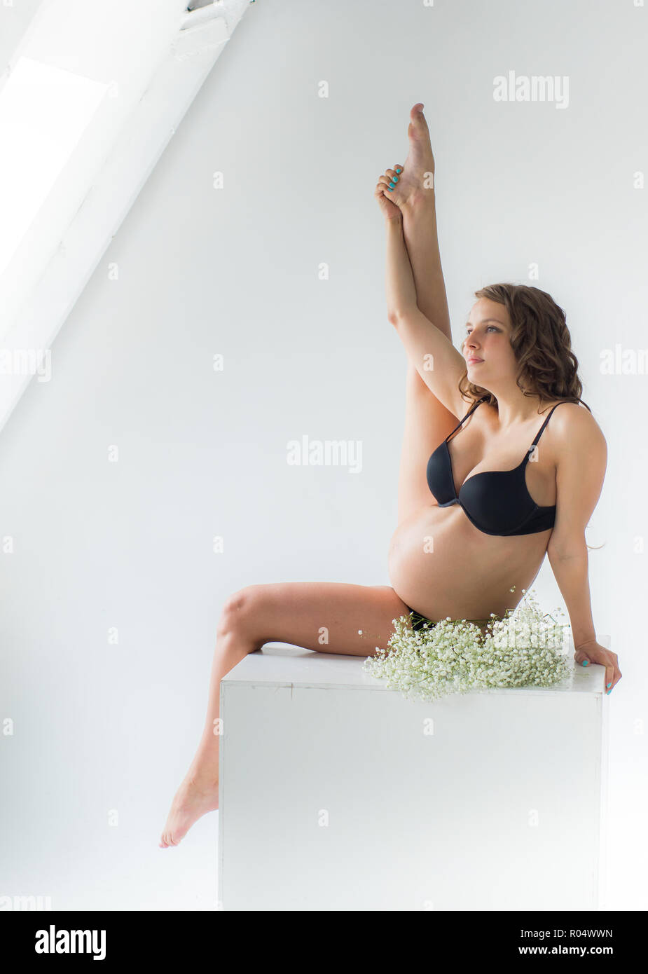 Seduto sul cubo grande donna incinta con mano e gamba in alto muro bianco su sfondo Foto Stock