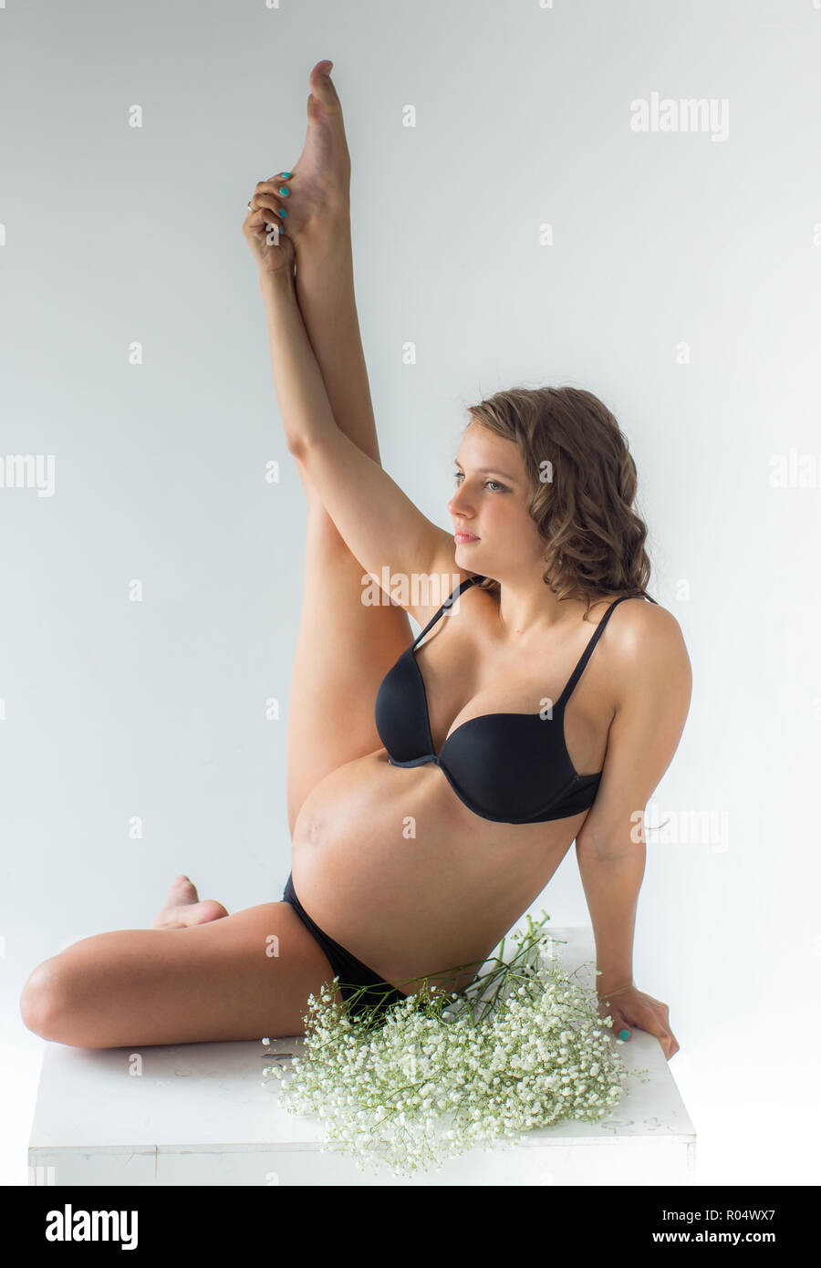 Seduto sul cubo grande donna incinta con mano e gamba in alto muro bianco su sfondo Foto Stock