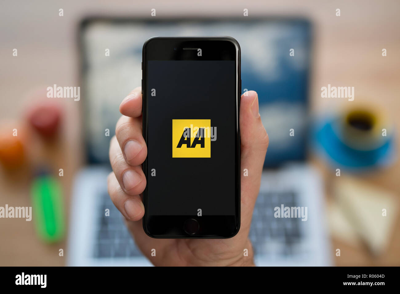Un uomo guarda al suo iPhone che visualizza il logo di AA, mentre se ne sta seduto alla sua scrivania per computer (solo uso editoriale). Foto Stock