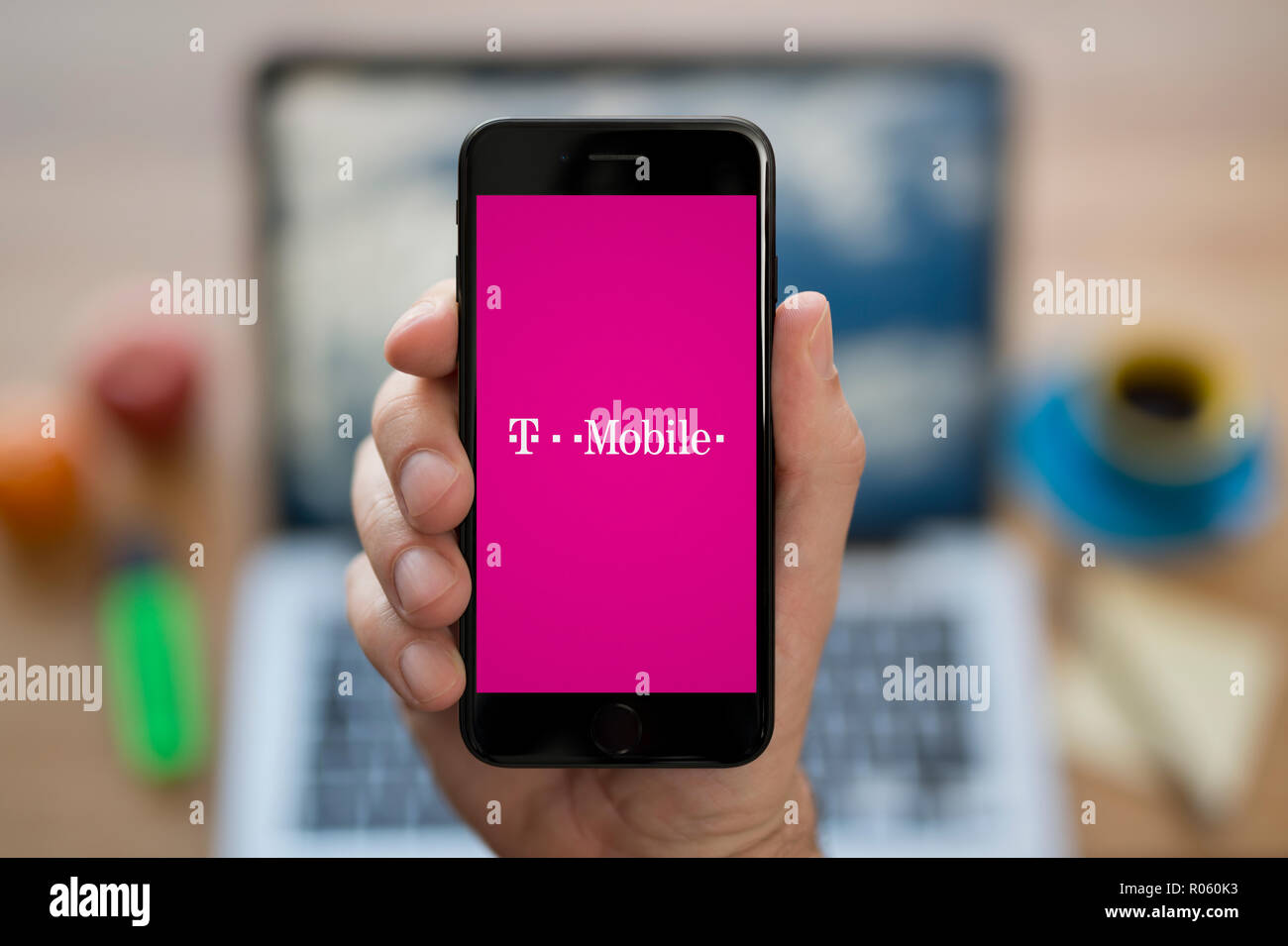 Un uomo guarda al suo iPhone che visualizza la T Mobile logo, mentre se ne sta seduto alla sua scrivania per computer (solo uso editoriale). Foto Stock