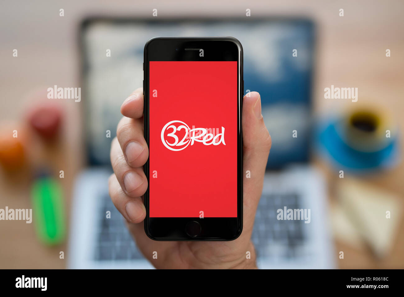 Un uomo guarda al suo iPhone che visualizza il 32logo rosso, mentre se ne sta seduto alla sua scrivania per computer (solo uso editoriale). Foto Stock