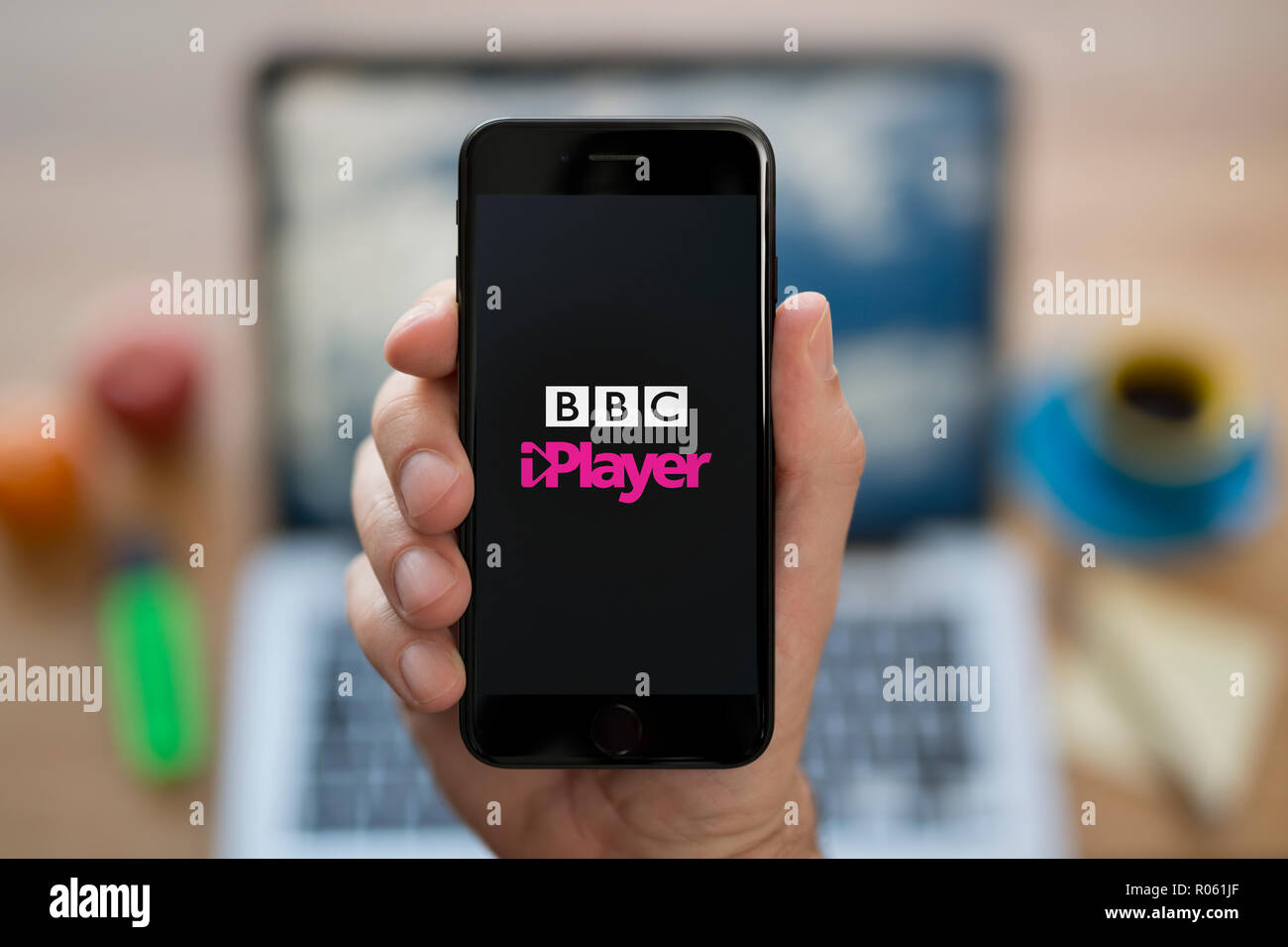 Un uomo guarda al suo iPhone che visualizza la BBC iPlayer logo, mentre se ne sta seduto alla sua scrivania per computer (solo uso editoriale). Foto Stock