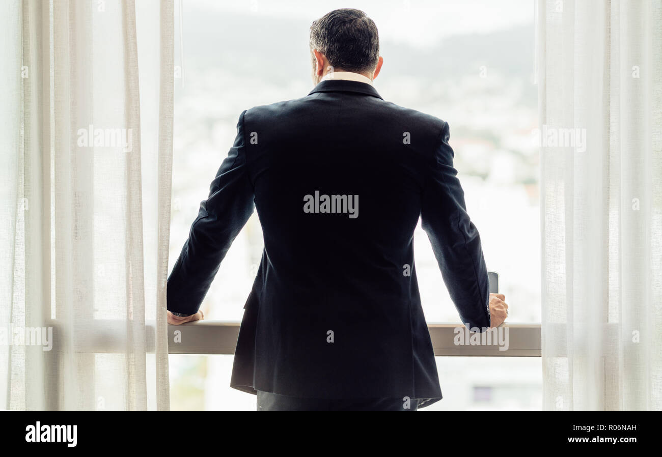 Vista posteriore di imprenditore in piedi da hotel finestra della camera. Uomo in business suit guardando fuori dalla finestra. Foto Stock
