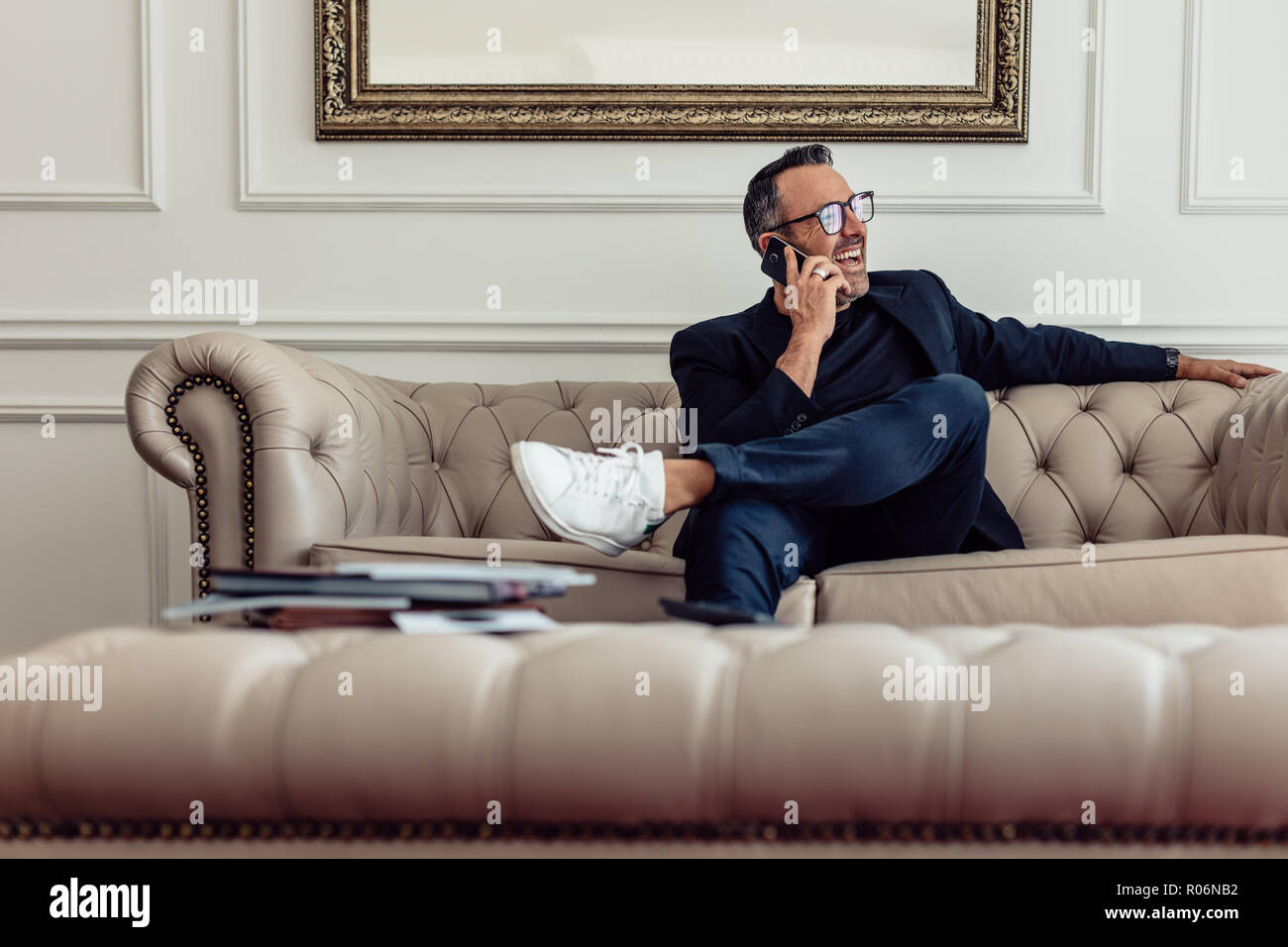 Felice adulto medio imprenditore parlando sul suo smart phone in camera d'albergo. Uomo in business suit seduti sul divano a parlare al telefono e guardando lontano sorridente Foto Stock