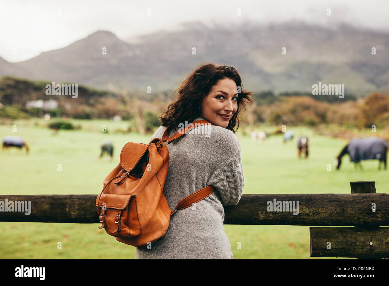 Vista posteriore della donna in piedi da staccionata in legno e guardando indietro alla fotocamera con i cavalli in background. Femmina da permanente ranch recinto. Foto Stock