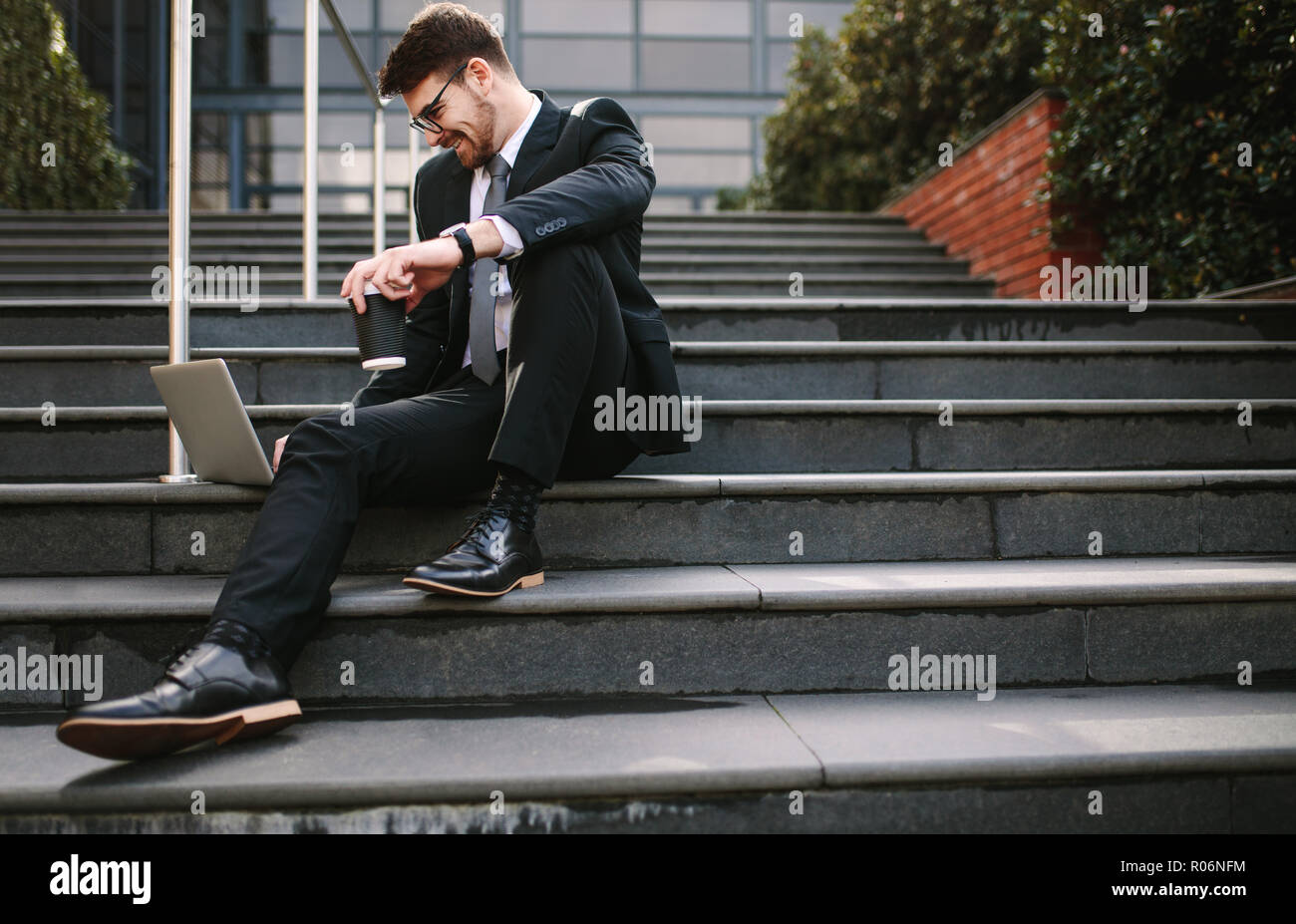 Sorridente azienda giovane uomo seduto sui gradini con laptop e caffè. Imprenditore rilassati guardando il laptop e sorridente mentre è seduto sui gradini al di fuori Foto Stock