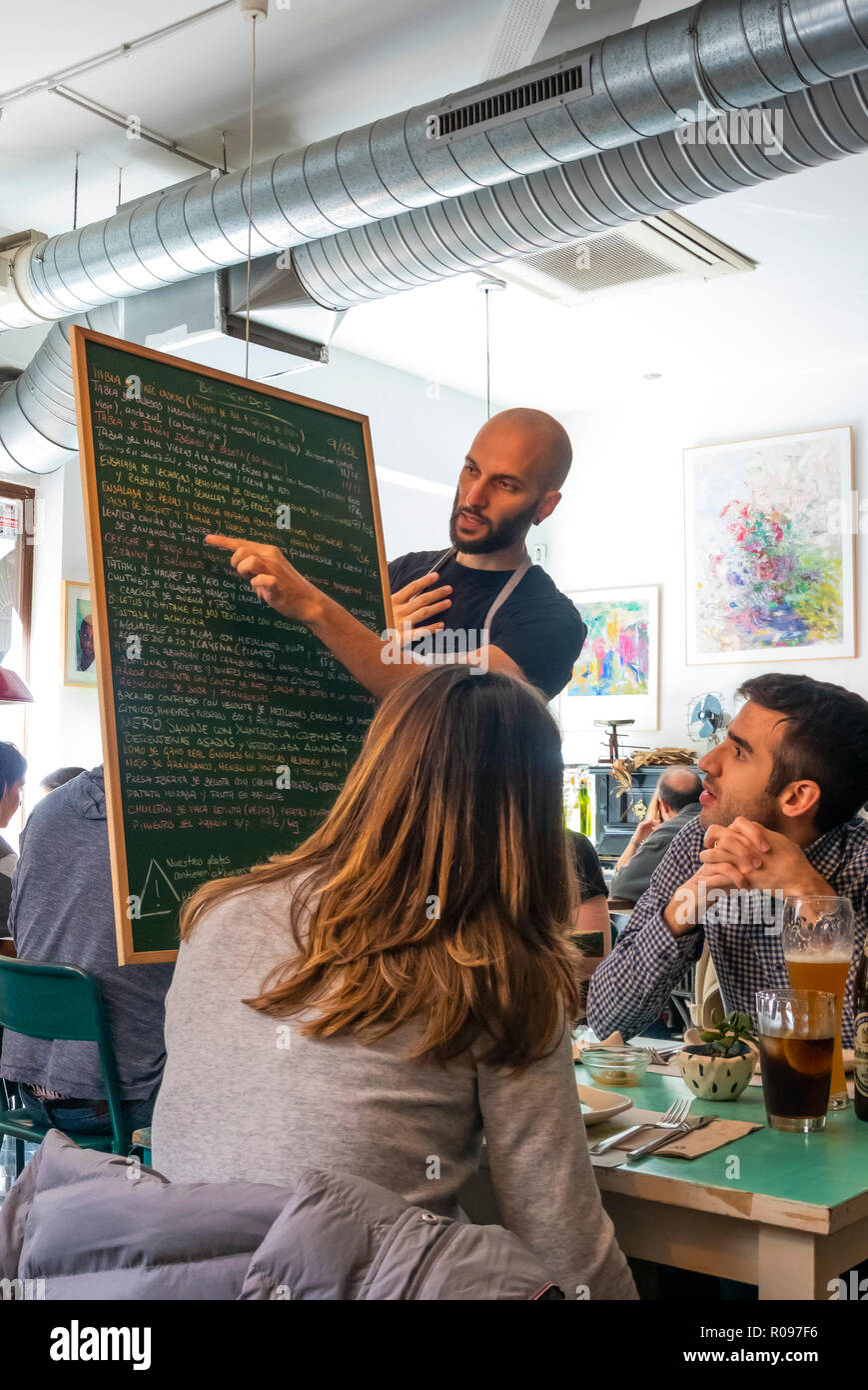 Un cameriere a Contenedor Ristorante Slow Food spiega gli elementi sulla lavagna giornalmente menu per una giovane coppia a Siviglia, Spagna Foto Stock