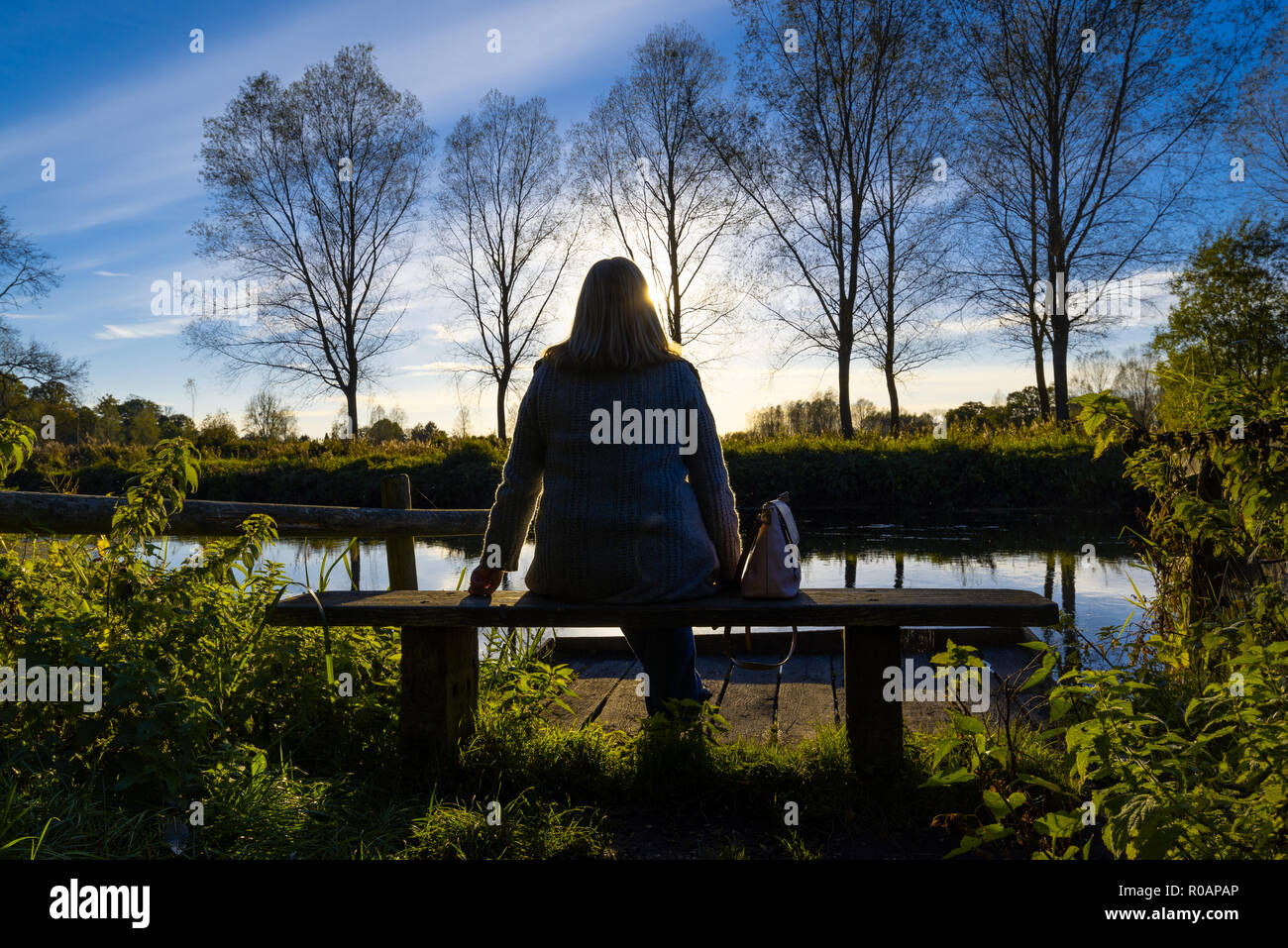 Donna seduta su una panchina di una tranquil riverside scena con un cielo blu riflessa nell'acqua e alberi e lussureggianti cespugli di rivestimento del lungofiume. Foto Stock