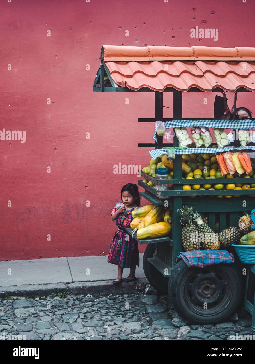 Piccolo giovane ragazza guatemalteco nel tradizionale abito Maya nascondersi dietro la sua famiglia di frutta carrello bancarella di strada Foto Stock