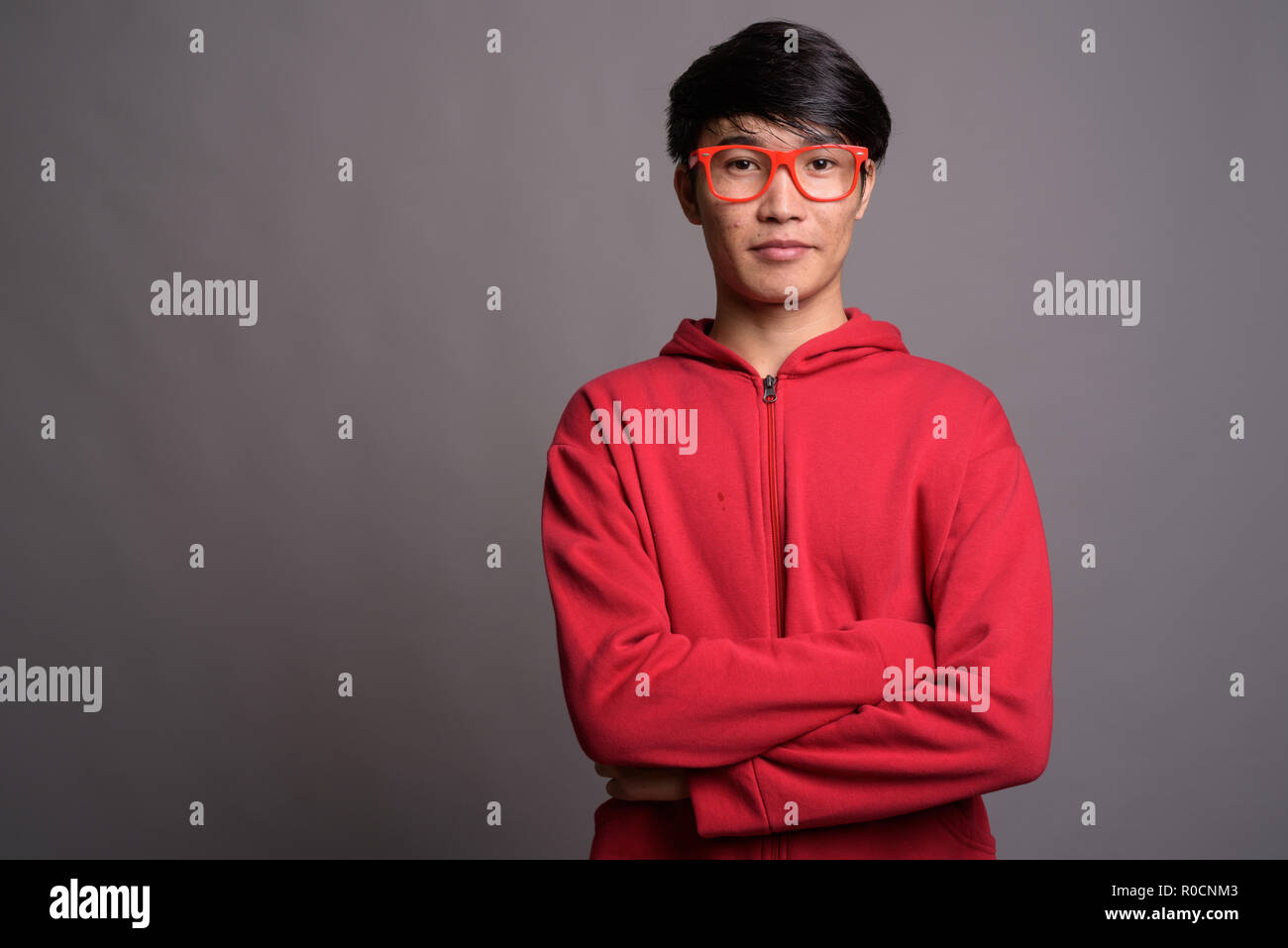 Giovane uomo asiatico indossando giacca rossa con occhiali contro il grigio Foto Stock