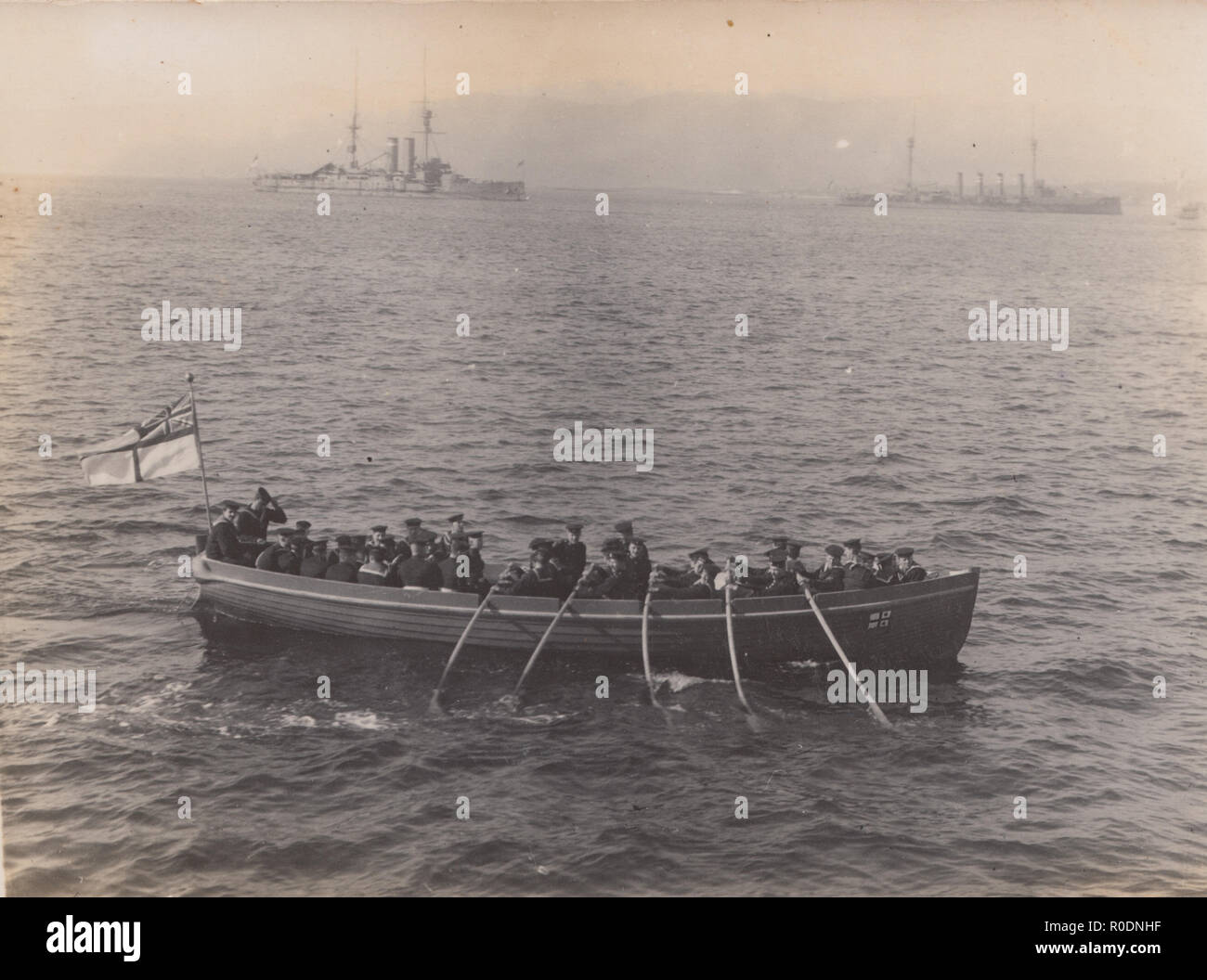 Vintage Cartolina fotografica di ?.B.S regata, Colonsay. Barca a remi di marinai con navi da guerra in background. Foto Stock
