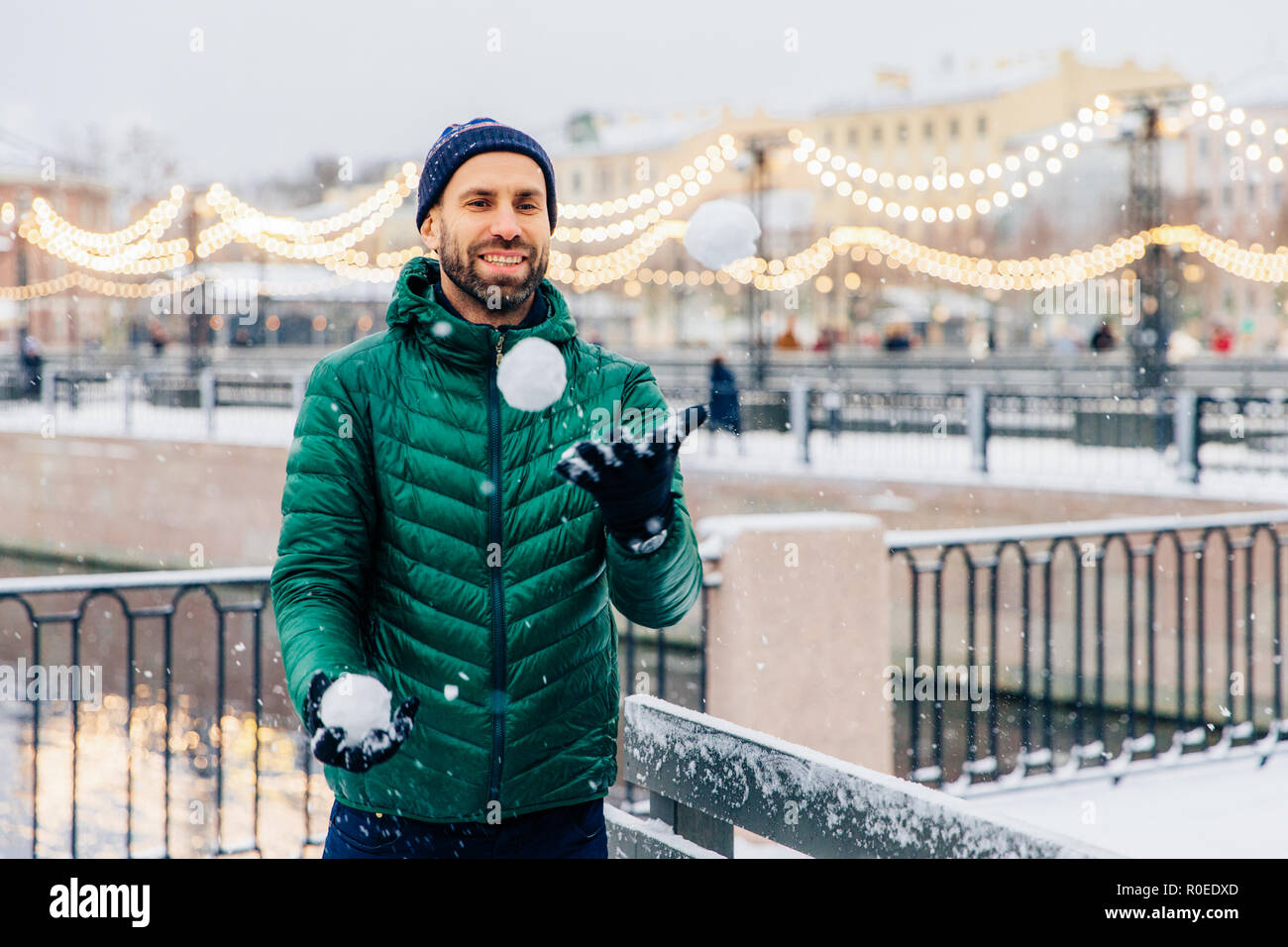 Colpo all'aperto di sorridenti barbuto maschio in una giacca calda e hat gioca snowballs come si erge contro belle luci, essendo di buon umore è gioioso e Foto Stock