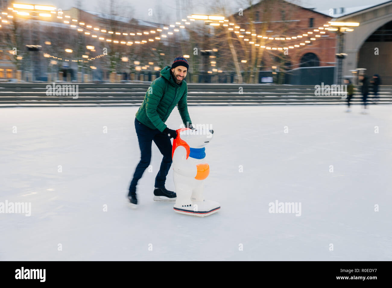 Foto del pattinatore maschio mantiene l'equilibrio sul pattino anello, sorge su pattini con aiuto di aiuto di skate, trascorre le domeniche invernali con divertimento, gode di frosty weathe nevoso Foto Stock