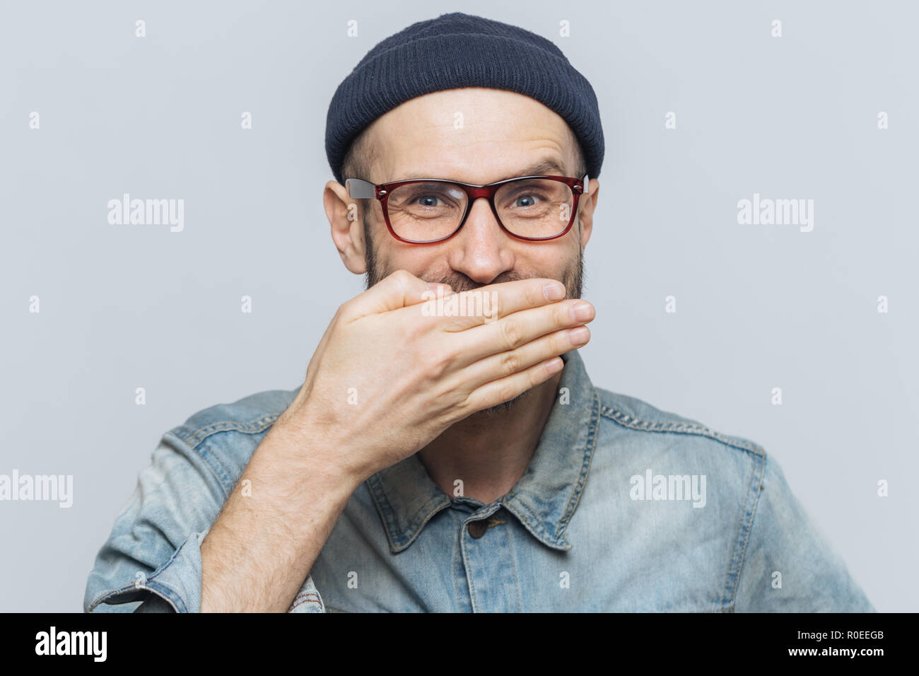 Funny Man con look accattivante giggles o ride con gioia, copre la bocca con la mano, cerca di fermare i sentimenti, sente aneddoto da amico, indossa gli occhiali, Foto Stock