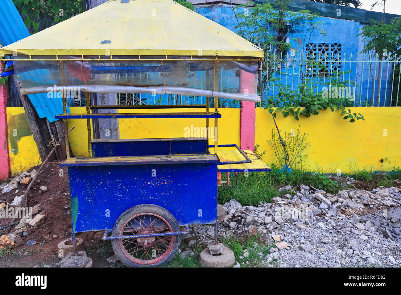 Abbandonato foodcart dipinte di giallo e blu accanto al giallo muro in cemento armato di una casa blu con reticolo metallico recinto sul lato di una strada nel cen Foto Stock