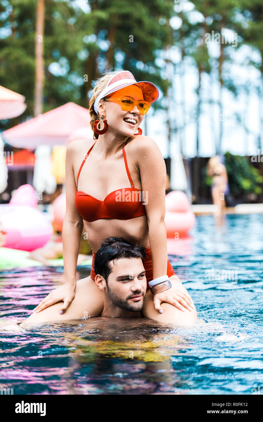 Donna sorridente in costume rosso che guarda lontano mentre è seduto su fidanzati spalle in piscina Foto Stock