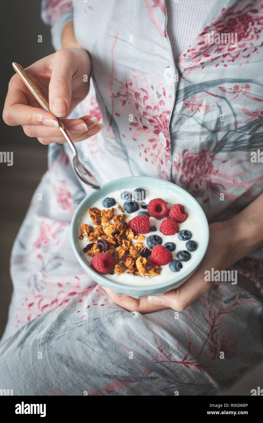 Irriconoscibile femmina in pijama mangiare yogurt con muesli, mirtilli e lamponi. Concetto di colazione vegetariana, mangiare sano, vita sana Foto Stock