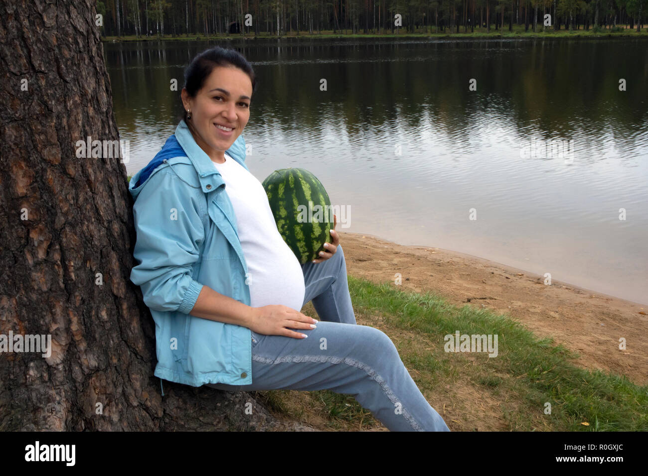 Attraente incinta brunette con anguria in posa vicino alla foresta calmo lago Foto Stock