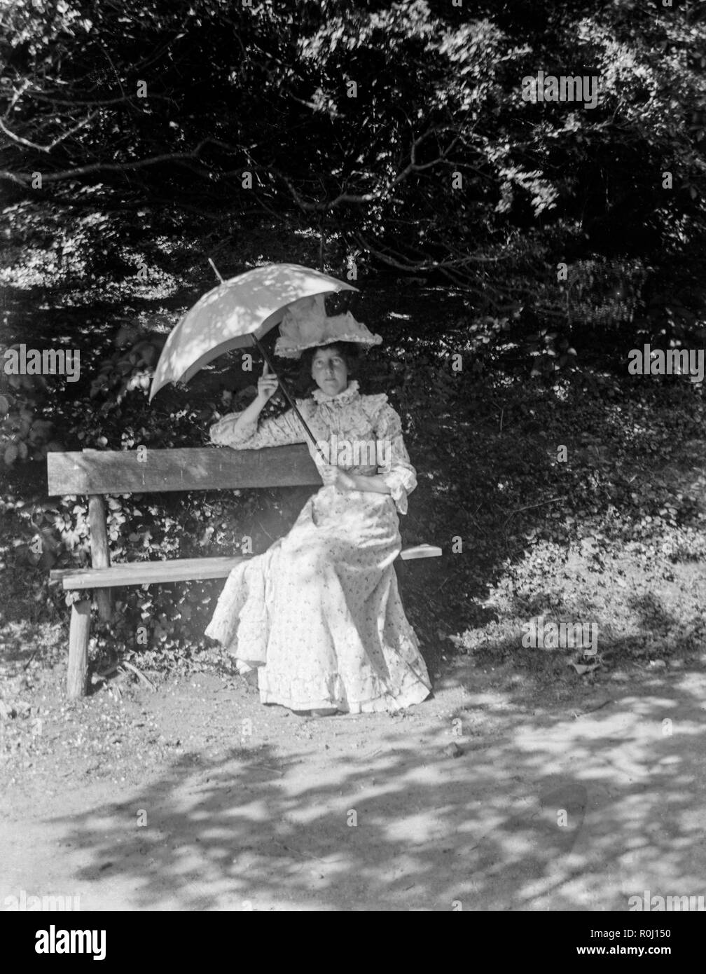 Un Edwardian fotografia in bianco e nero che mostra una giovane donna seduta su una panchina sotto un albero, vestito in belle abiti formali, mentre tiene un ombrellone sopra la sua testa. Foto Stock