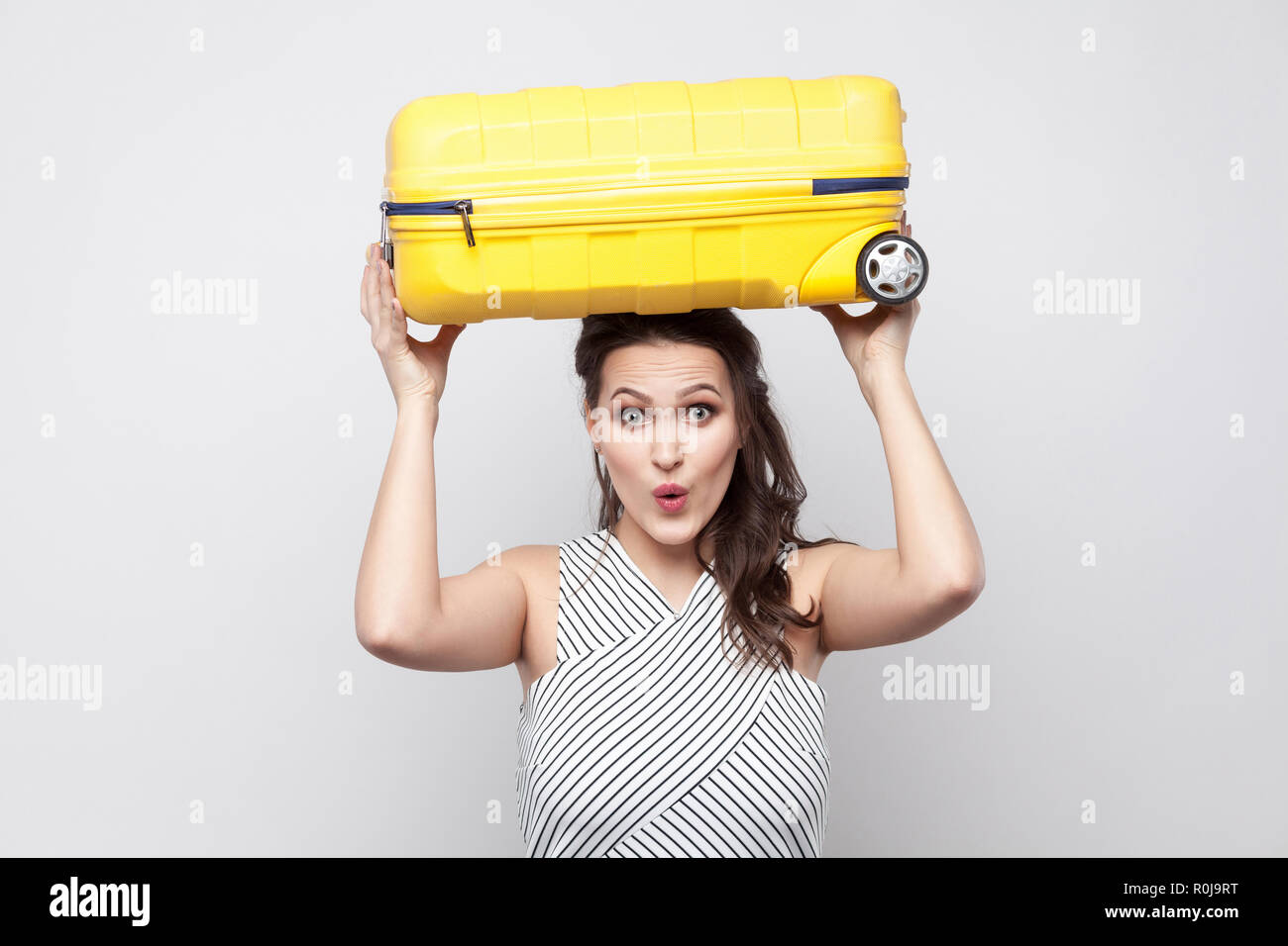 Funny forte giovane viaggiatore donna in abito a righe in piedi e tenere sopra la testa gialla con valigia shoked faccia e pronto per il viaggio. Stu Indoor Foto Stock