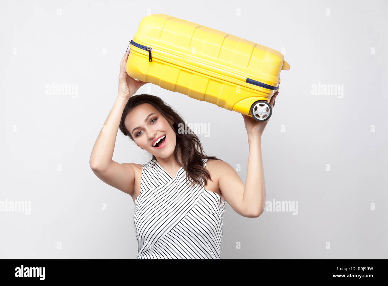 Funny forte giovane viaggiatore donna in abito a righe in piedi e tenere sopra la testa gialla valigia, sorriso toothy e pronto per le vacanze. Piscina San Foto Stock