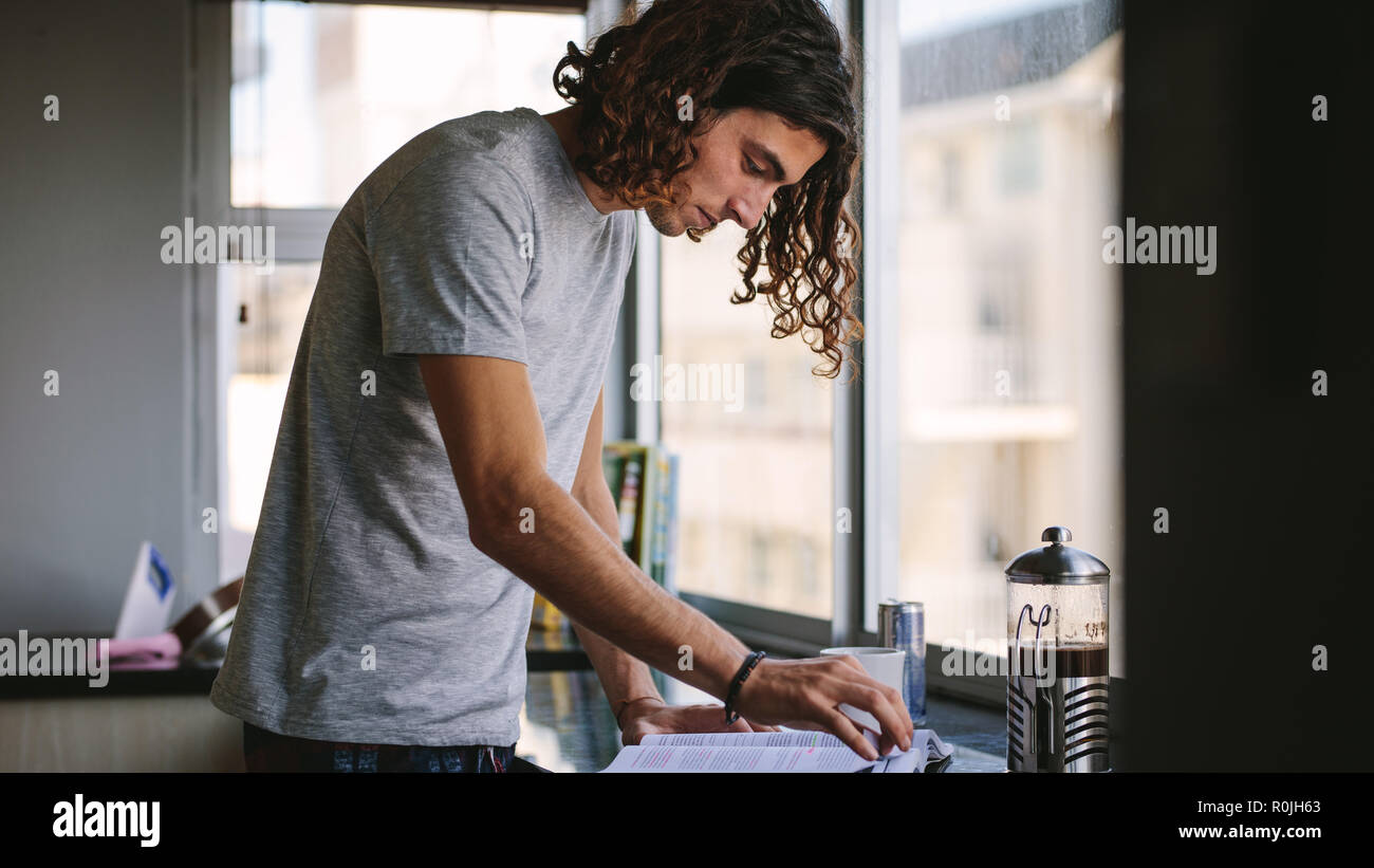 Giovane uomo in piedi in cucina accanto a una finestra e la lettura di un libro. Studente per l'esame durante la preparazione del caffè in cucina. Foto Stock