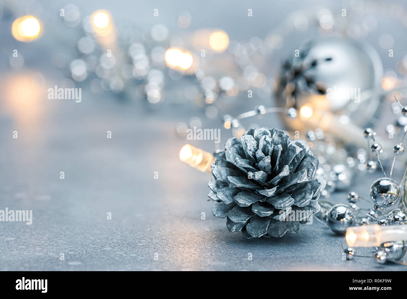 Incandescente le luci di Natale ghirlanda e cono di pino con jingle bell su sfocato sfondo color argento Foto Stock