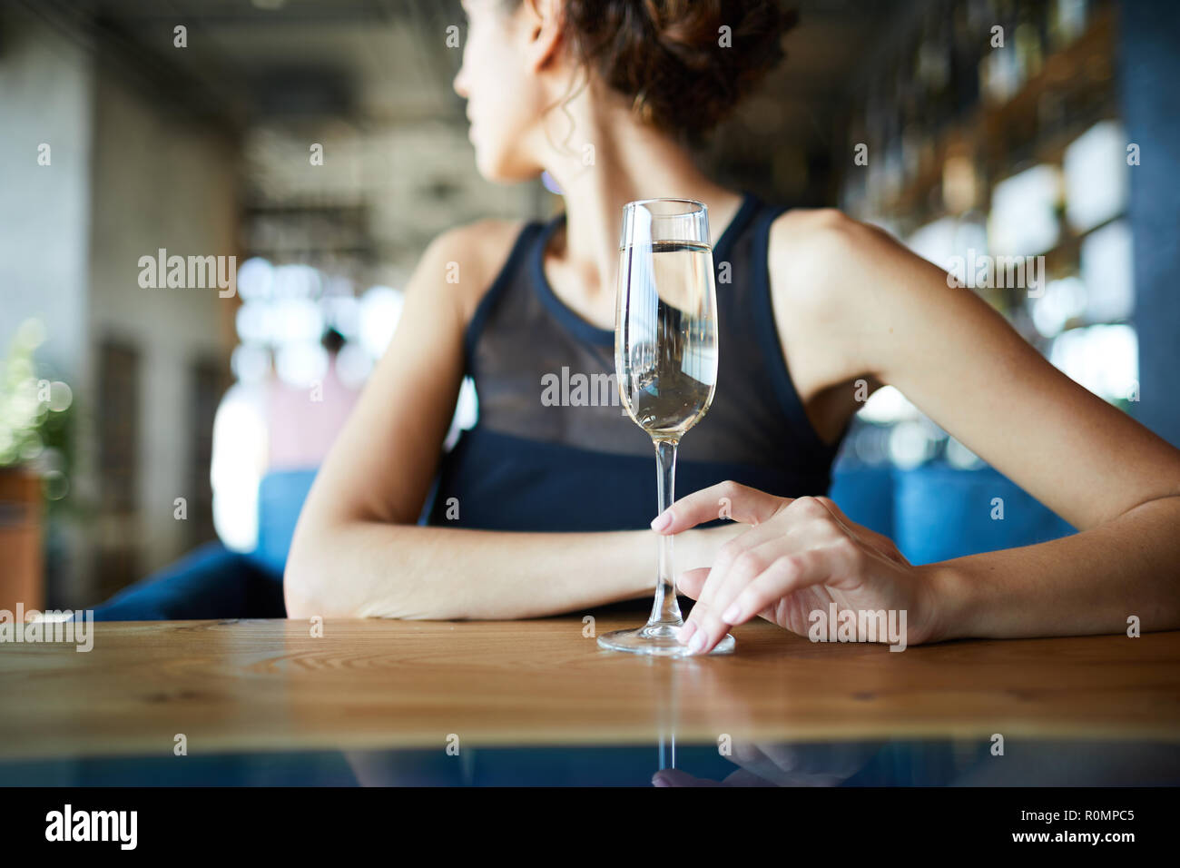 Giovane donna seduta al tavolo in cafe e toccando flûte di champagne mentre guardando sopra la sua spalla Foto Stock