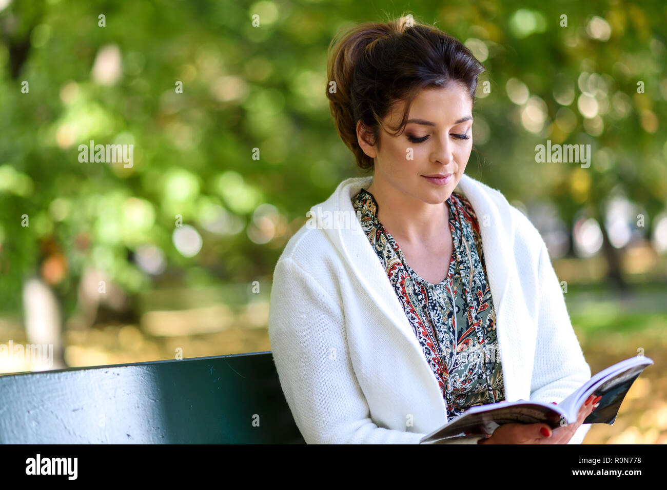 Bella giovane donna seduta su una panchina nel parco e la lettura di un libro su una soleggiata giornata autunnale. Foto Stock