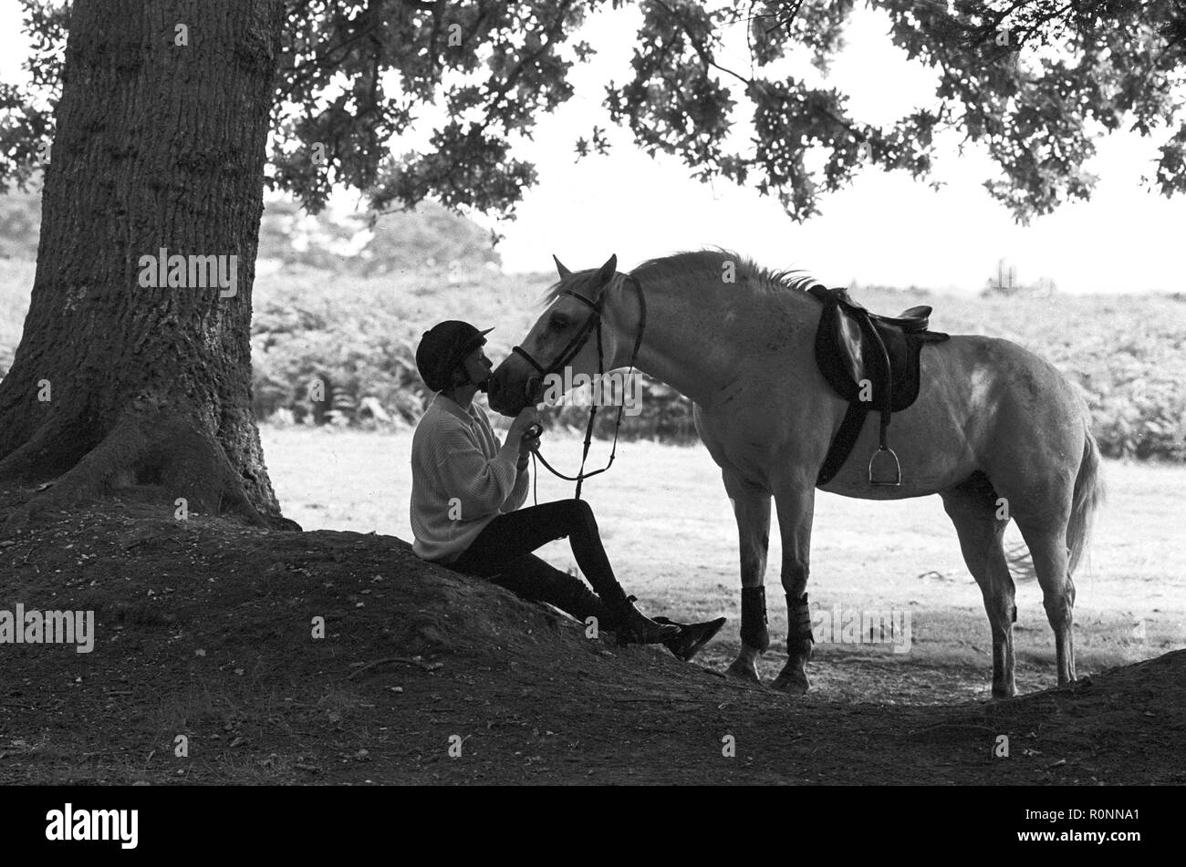 Una giovane ragazza in equipaggiamento da guida si trova sotto un albero estivo nella New Forest, Hampshire, e dà il suo pony bianco e migliore amico un bacio. Fotografia b&w vintage Foto Stock
