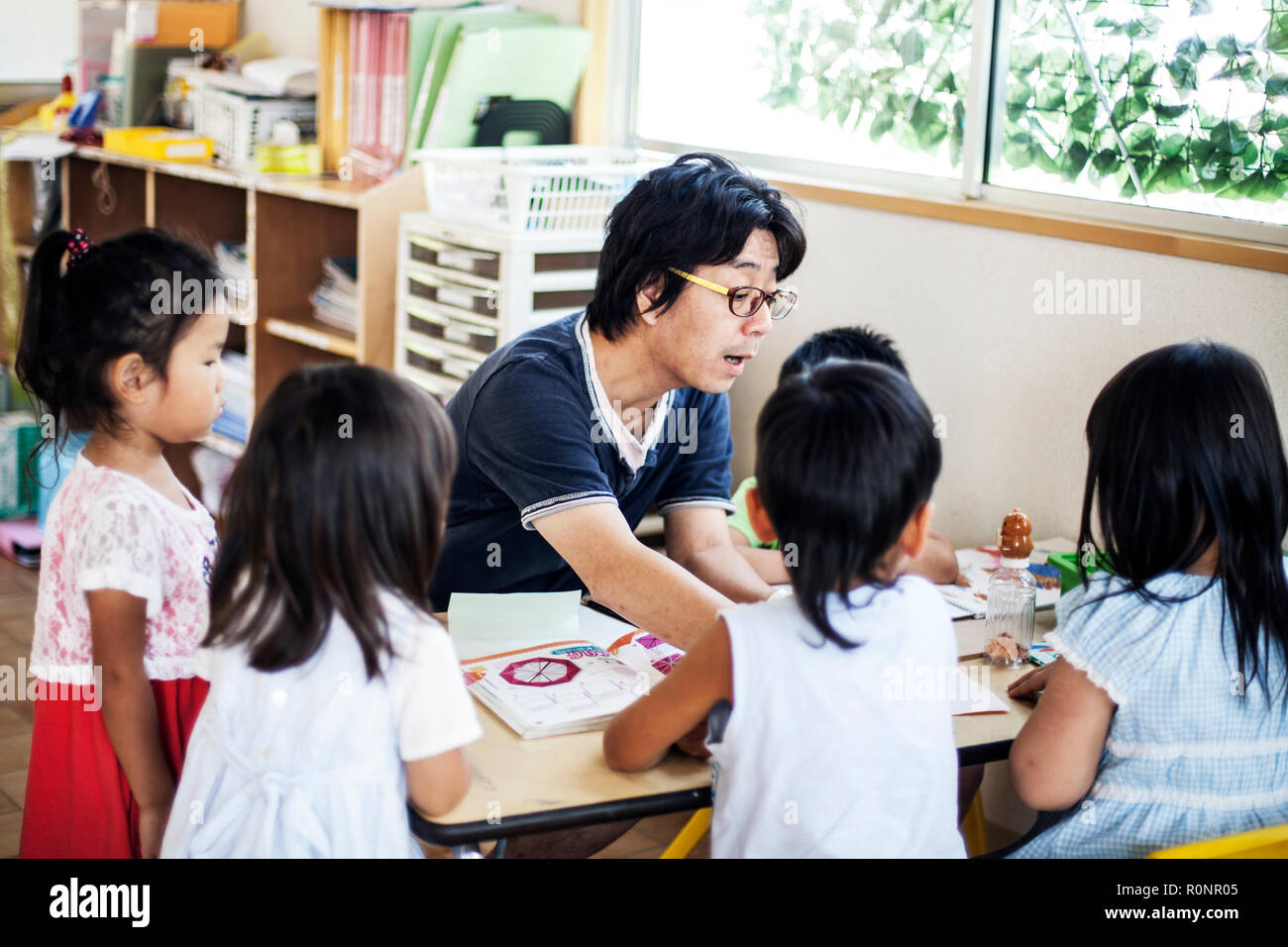 Insegnante di sesso maschile di parlare a un gruppo di bambini in una tabella n un giapponese di età prescolare. Foto Stock