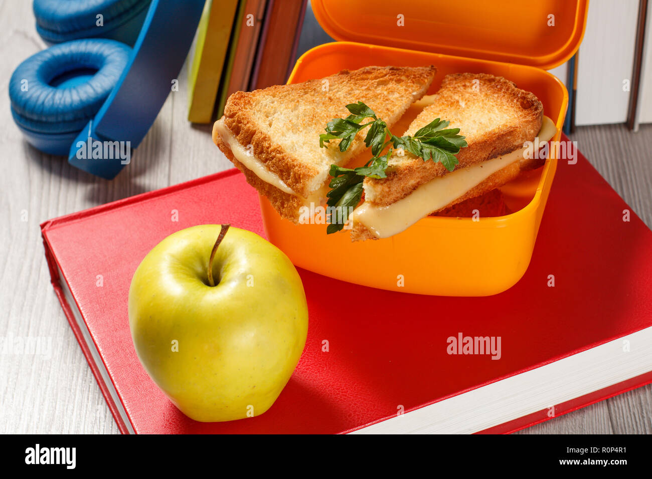 Giallo casella a sandwich con le fette biscottate di pane, formaggio e prezzemolo verde, verde mela, cuffie e rigida libri sullo sfondo. Rottura della scuola Foto Stock