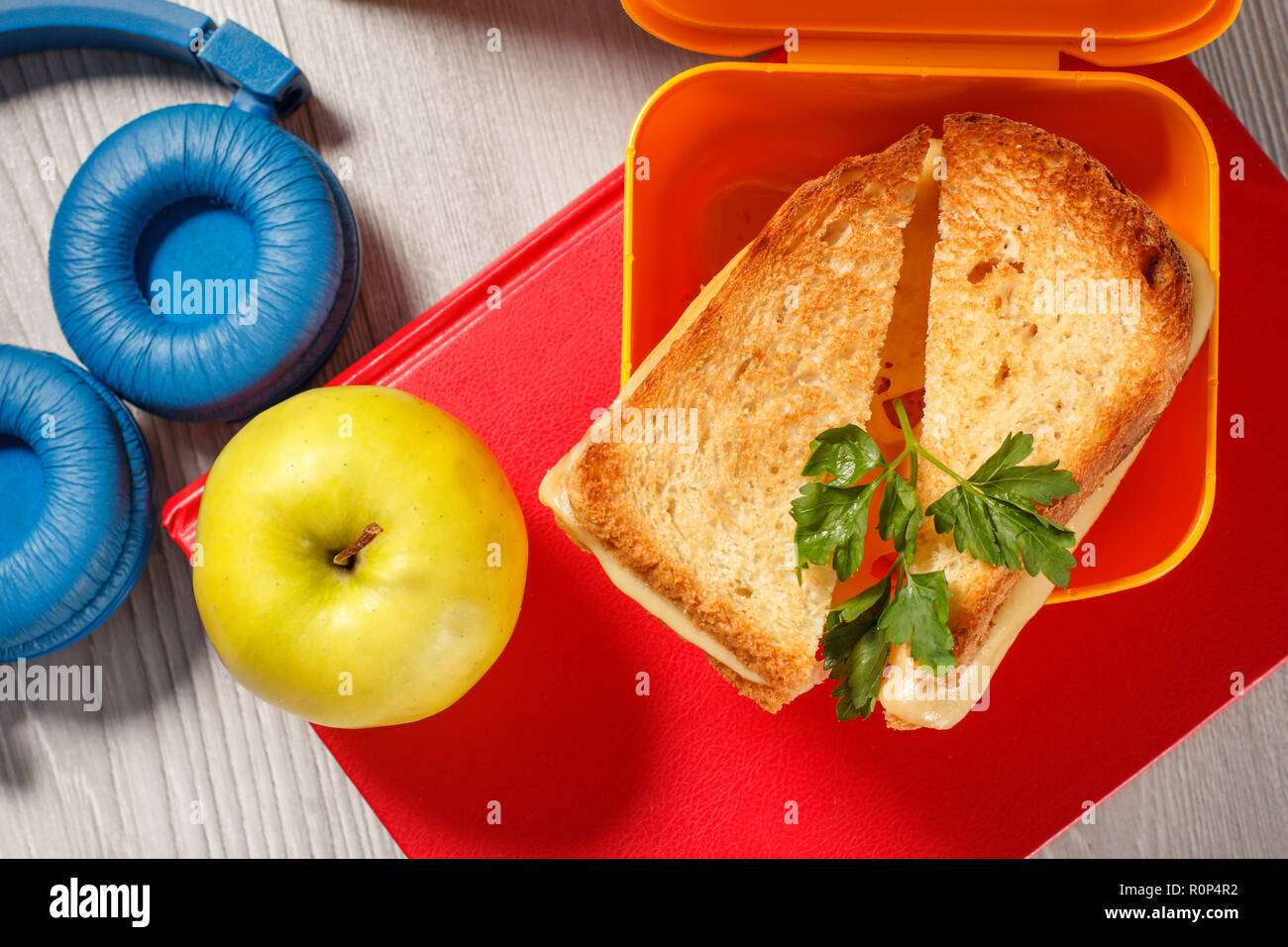 Giallo casella a sandwich con le fette biscottate di pane, formaggio e prezzemolo verde, verde mela, cuffie e rigida libri sullo sfondo. Rottura della scuola Foto Stock