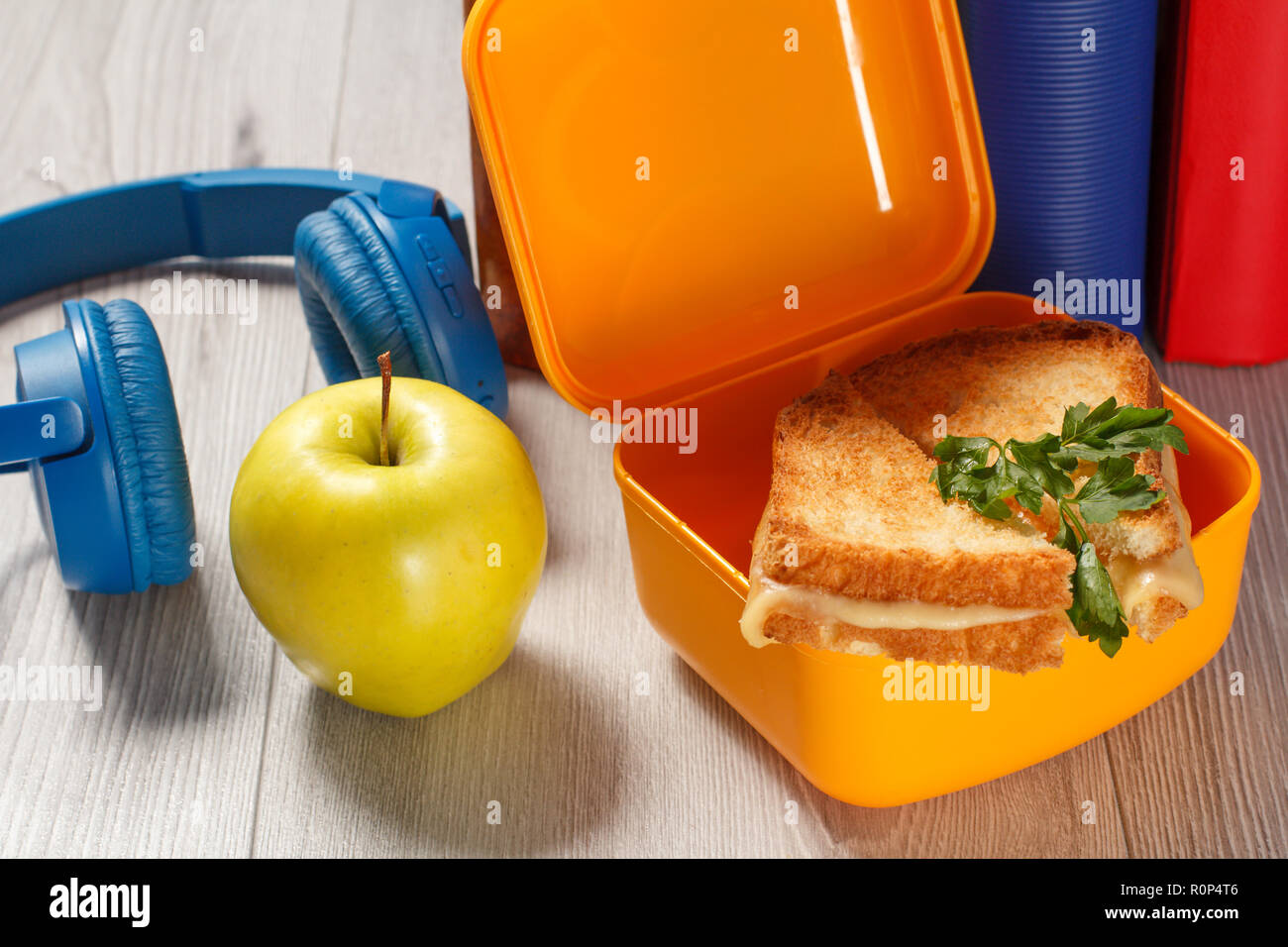 Giallo casella a sandwich con le fette biscottate di pane, formaggio e prezzemolo verde, verde mela, cuffie e rigida libri sullo sfondo. Rottura della scuola Foto Stock