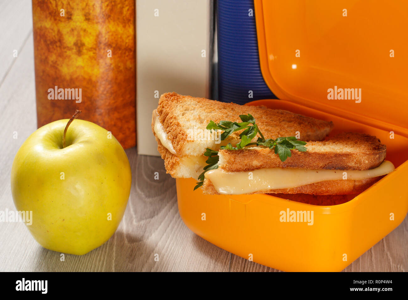 Giallo casella a sandwich con le fette biscottate di pane, formaggio e prezzemolo verde, verde mela e rigida libri sullo sfondo. La prima colazione a scuola. Foto Stock