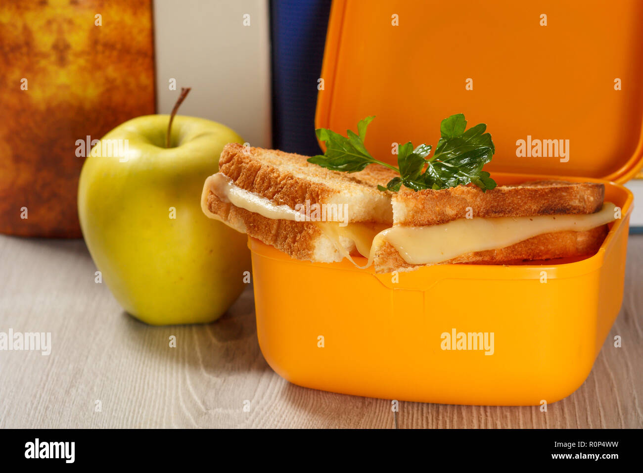 Giallo casella a sandwich con le fette biscottate di pane, formaggio e prezzemolo verde, verde mela e rigida libri sullo sfondo. La prima colazione a scuola. Foto Stock