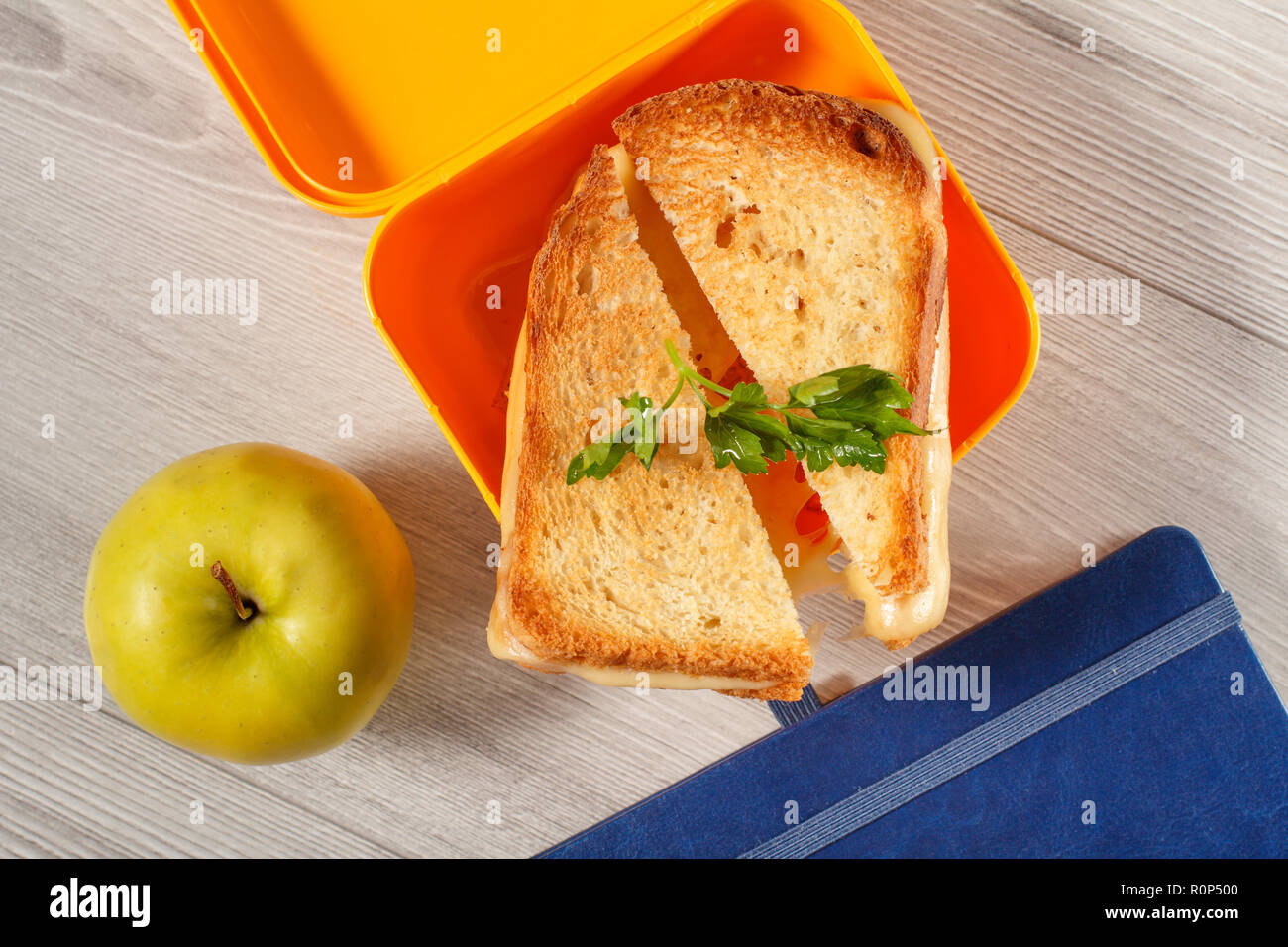 Giallo casella a sandwich con le fette biscottate di pane, formaggio e prezzemolo verde, verde mela e notebook su scrivania in legno. Vista dall'alto. La prima colazione a scuola. Foto Stock
