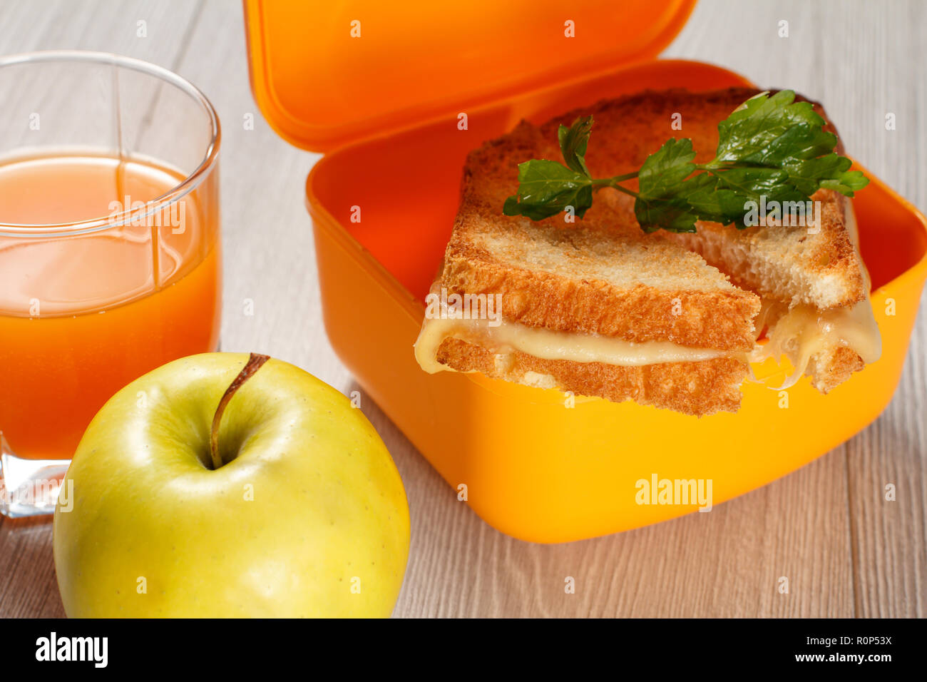 Giallo casella a sandwich con le fette biscottate di pane, formaggio e prezzemolo verde, verde mela e un bicchiere di succo di arancia sulla scrivania in legno. La prima colazione a scuola. Foto Stock