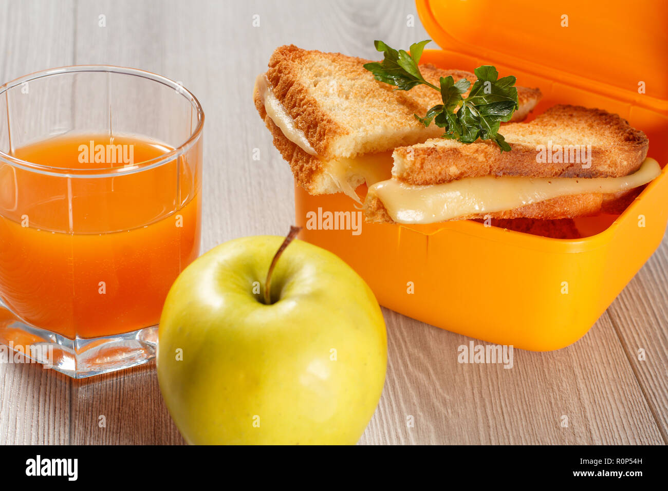 Giallo casella a sandwich con le fette biscottate di pane, formaggio e prezzemolo verde, verde mela e un bicchiere di succo di arancia sulla scrivania in legno. La prima colazione a scuola. Foto Stock