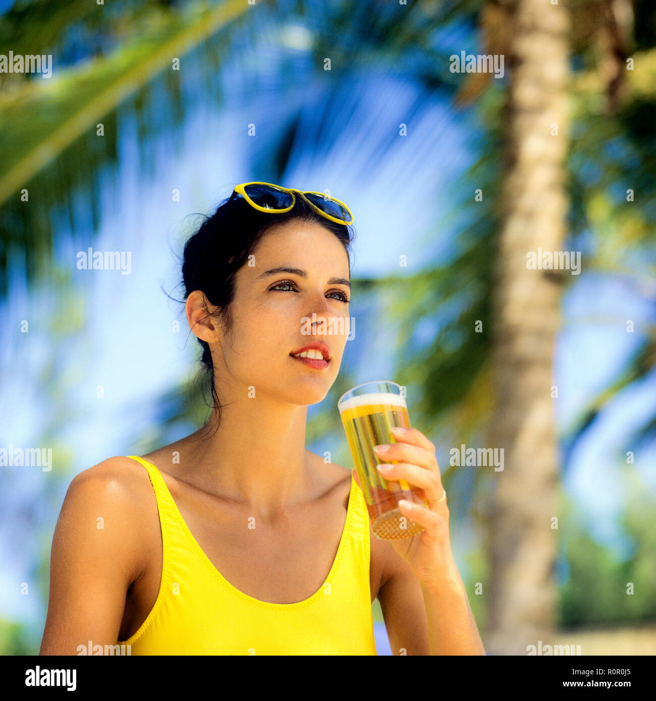 Giovane donna con il costume da bagno giallo tenendo un bicchiere di birra, palme sfondo, Guadalupa, French West Indies, Foto Stock