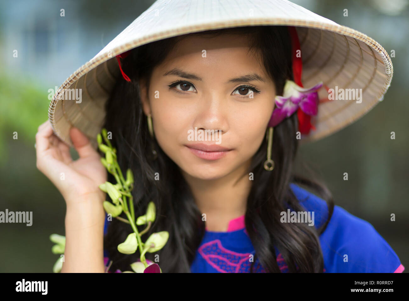 Attraente grave giovane donna vietnamita in una forma conica cappello di paglia con fiori freschi nei suoi capelli guardando la telecamera con la sua mano a rasamento Foto Stock