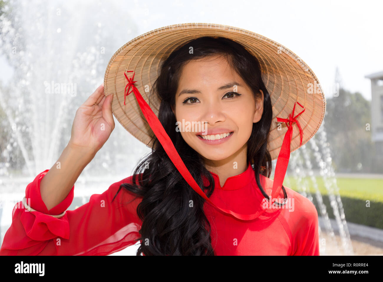 Donna vietnamita in un rosso Ao IAM e cappello conico in piedi davanti ad una fontana sorridente verso la telecamera Foto Stock