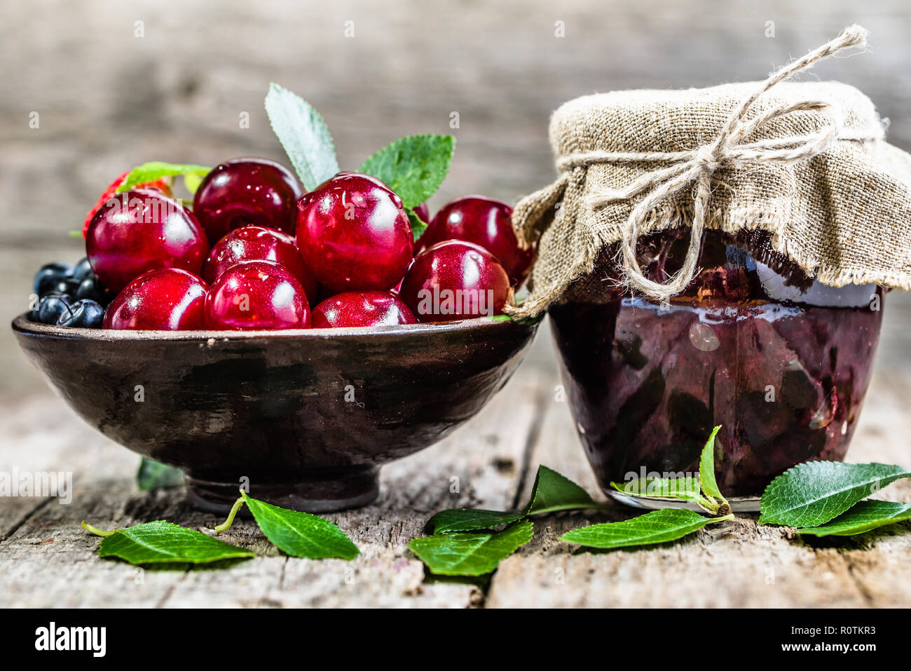 Confettura di ciliegie in vaso e le ciliege fresche in una ciotola, conserve fatte in casa su sfondo rustico Foto Stock