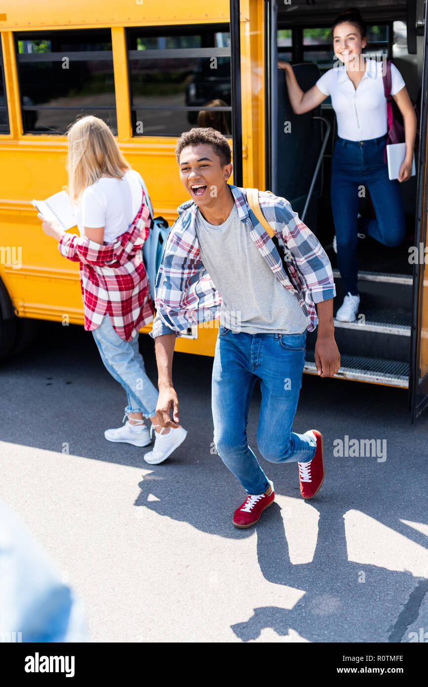 Ridendo African American schoolboy in esecuzione al di fuori della scuola bus con i compagni di scuola femminile Foto Stock