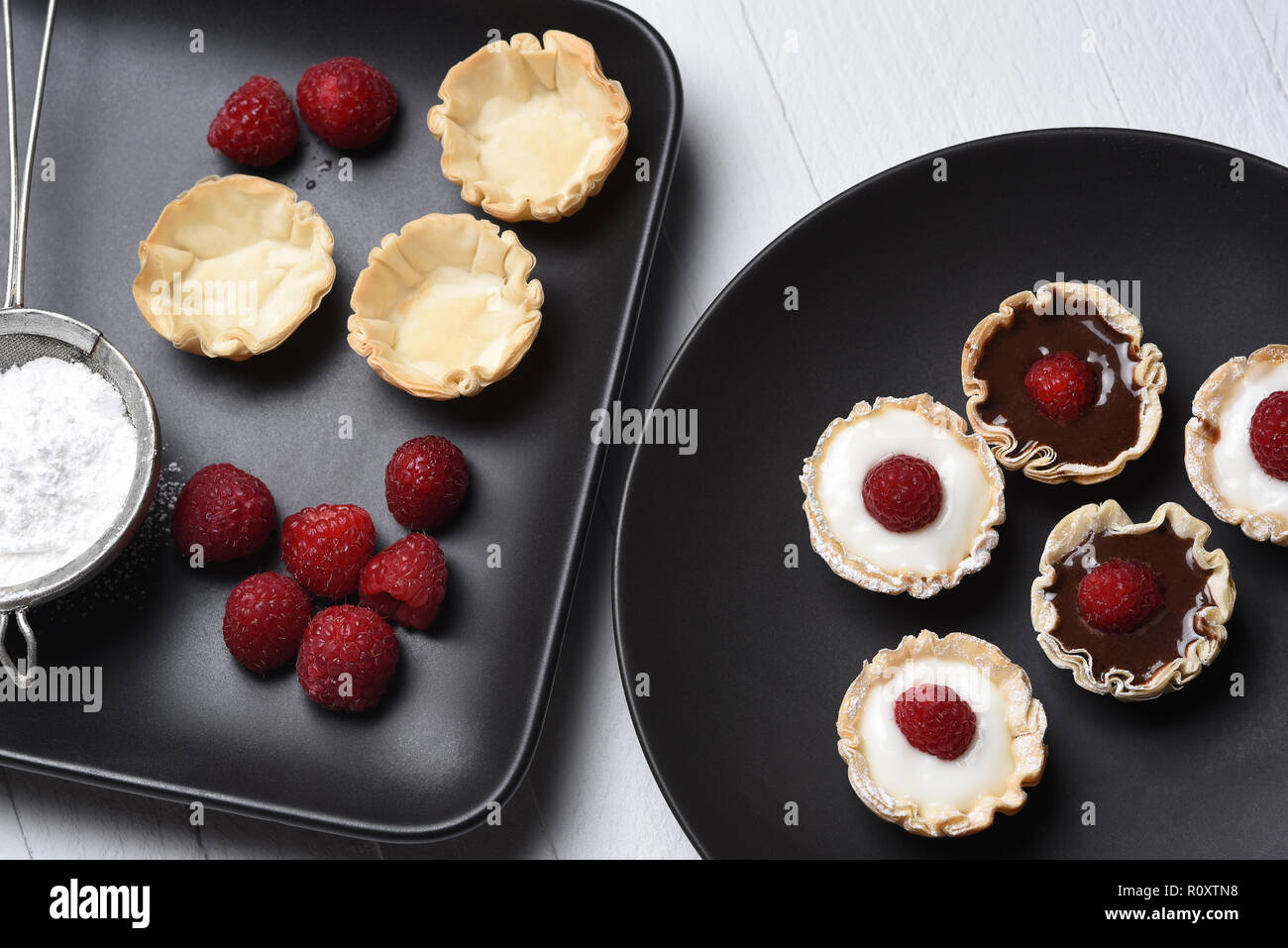 Lay piatto di cioccolato e crema alla vaniglia gusci di fillo e rabboccato con lamponi. Gli ingredienti su di una piastra e di dolci assemblato su un altro. Foto Stock