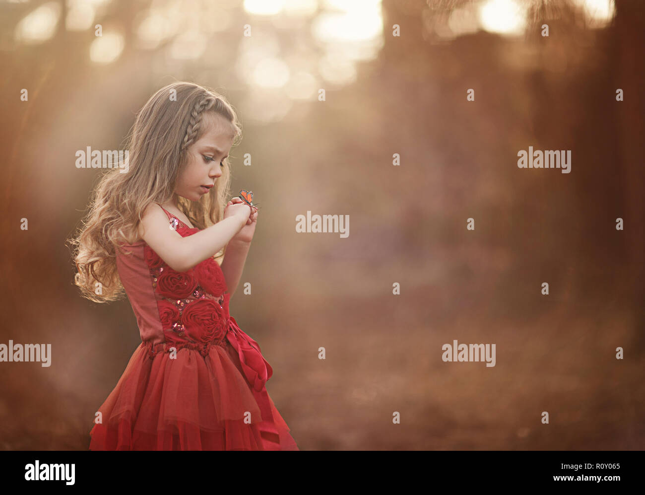 Giovane ragazza in un bel vestito rosso sta cercando in un timore reverenziale per una farfalla che ha atterrato sulla sua mano Foto Stock