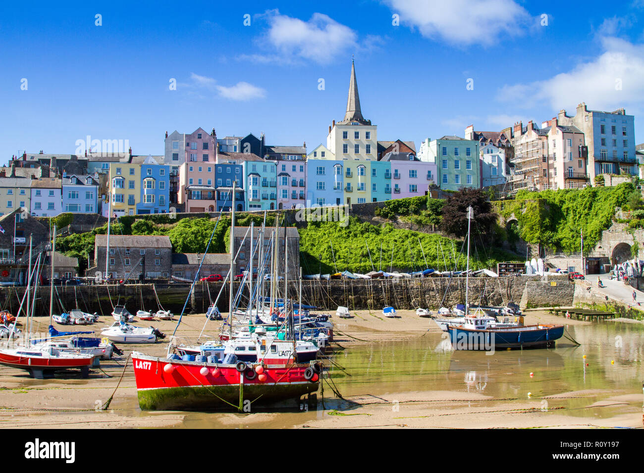 Bassa marea a Tenby, Pembrokeshire, Wales, Regno Unito. Barche a vela e yacht spiaggiata contro il dipinto di case a schiera e chiesa. Città di mare Foto Stock