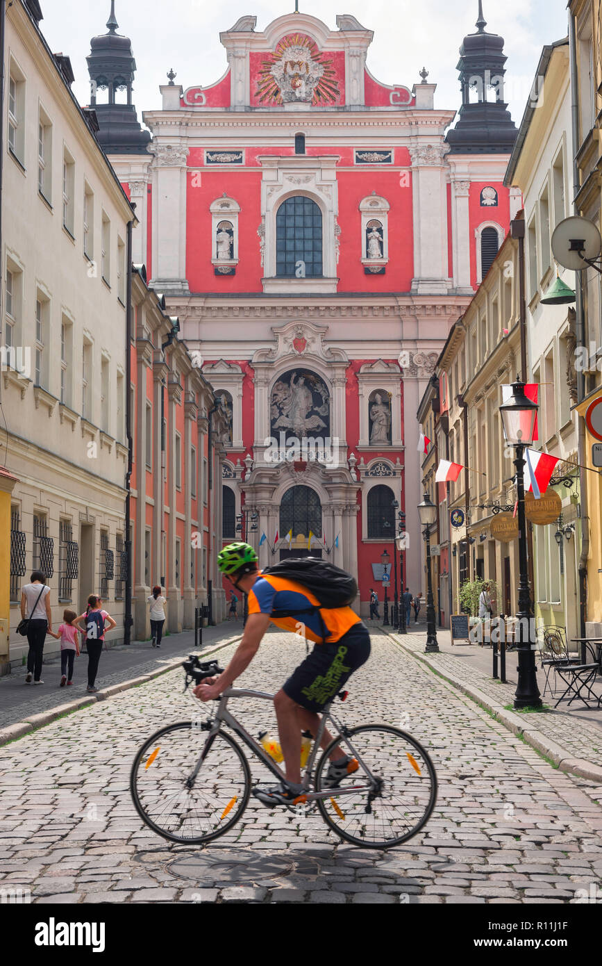 Viaggiare in bicicletta, un maschio ciclista in sella alla sua bicicletta attraverso Poznan Città Vecchia in Polonia sguardi alla facciata barocca della chiesa parrocchiale di San Stanislao. Foto Stock