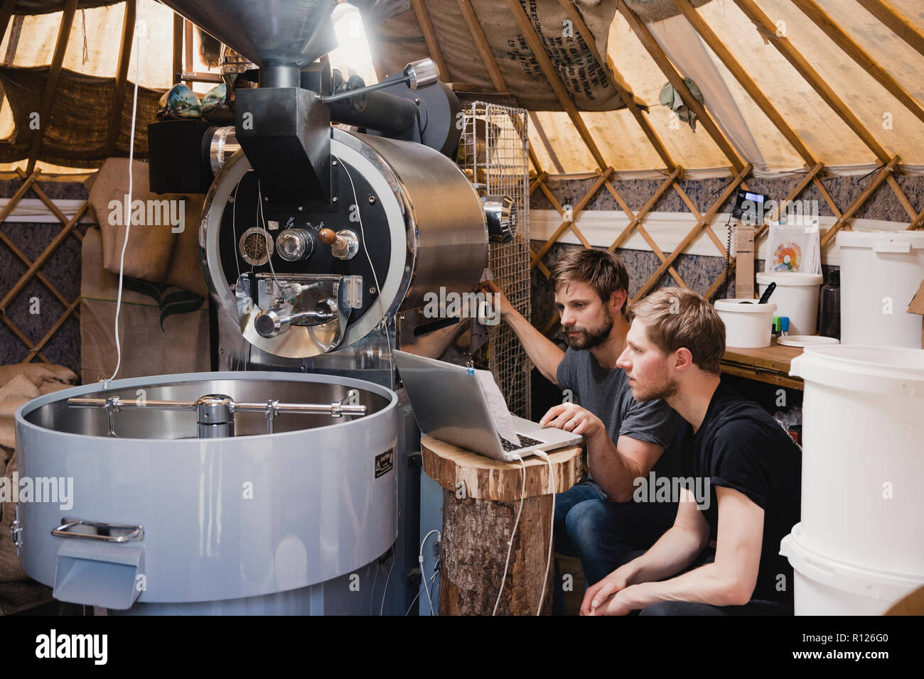 Due metà uomini adulti seduti in una stanza con un chicco di caffè tostatura di macchina. Stanno guardando il laptop e il fissaggio della macchina, assicurandosi che si tratta di un lavoro Foto Stock